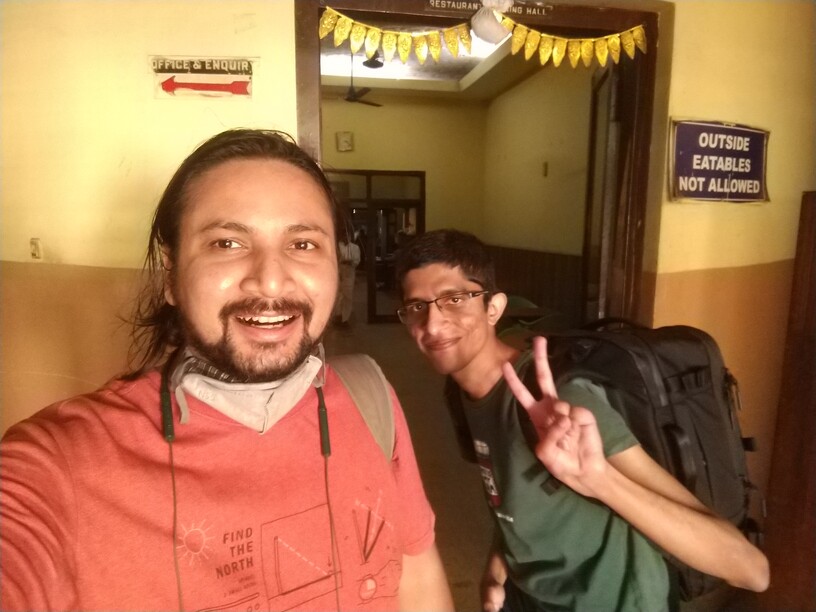 Selfie of two men outside a wall with a wooden doorway. Man on the left is Kashish, wearing an orange T-shirt. There are two masks pulled below his chin, and he has a Bluetooth headset around his neck. Man on the right is Vivek, wearing a green T-shirt and a black backpack, smiling and flashing a V-sign. The wall has signs saying "OFFICE & ENQUIR[Y]" to the left of the door and "OUTSIDE EATABLES NOT ALLOWED" to the right of the door.